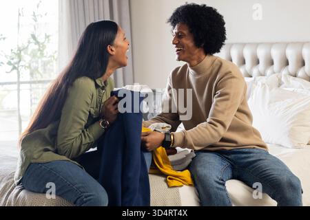 Rire divers couple pliant le linge sur le lit dans une chambre à coucher légèrement éclairée, avec panier à linge Banque D'Images