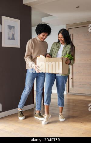 Inspecter la boîte d'épicerie en carton, couple diversifié debout dans l'entrée de la maison avec du pain et des légumes verts Banque D'Images
