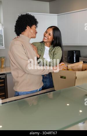 Souriant couple diversifié déballage boîte en carton de produits d'épicerie sur l'îlot de cuisine moderne, avec des fruits Banque D'Images