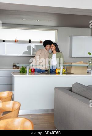 Couple souriant diversifié appuyé contre le garde-corps en verre à l'îlot de cuisine, avec des épiceries fraîches Banque D'Images