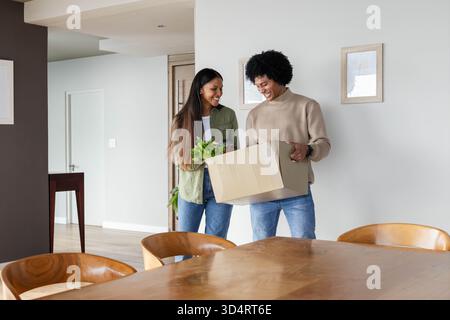 Déballage d'un couple diversifié tenant une boîte en carton avec des verts feuillus frais dans la salle à manger moderne Banque D'Images