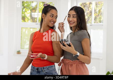 Application de maquillage diverses amies féminines debout par la fenêtre à la maison, avec rouge compact, palette noire Banque D'Images