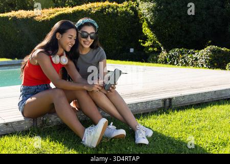 Souriant diverses amies féminines se penchant sur un smartphone sur la terrasse de la piscine, avec des écouteurs et des lunettes de soleil Banque D'Images