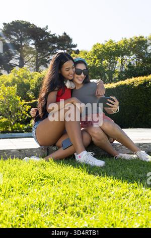 Souriant diverses amies féminines appuyées ensemble sur un banc en bois dans le parc, avec smartphone Banque D'Images