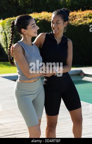 Embrasser diverses amies féminines debout sur la terrasse de la piscine en bois dans la cour arrière, avec smartwatch de fitness Banque D'Images