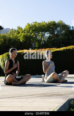 Méditer diverses amies féminines assises sur une terrasse en bois, avec des tapis de yoga et des écouteurs blancs Banque D'Images