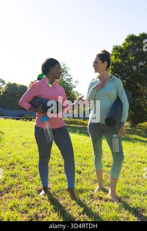 Bavardez diverses amies féminines tenant des tapis de yoga et des bouteilles d'eau sur un terrain herbeux avec des tapis noirs Banque D'Images