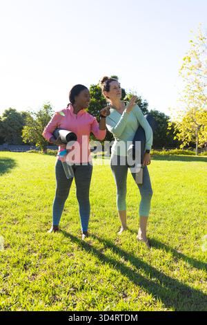 Marchant diverses amies féminines portant des tapis de yoga et une bouteille d'eau avec smartphone sur un terrain herbeux Banque D'Images