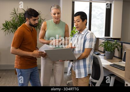 Discuter de divers collègues examinant des documents imprimés et pointant vers l'ordinateur portable dans un bureau ouvert Banque D'Images