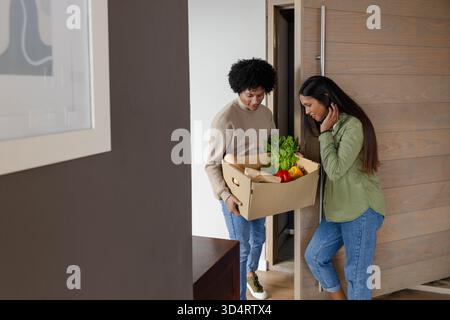 Couple diversifié échangeant boîte d'épicerie à l'entrée de la maison moderne, avec laitue et baguette Banque D'Images