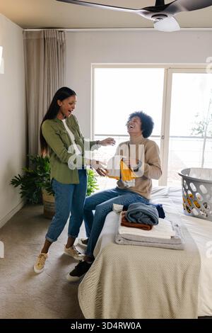 Rire couple diversifié plier le linge ensemble dans une chambre moderne, avec panier à linge Banque D'Images