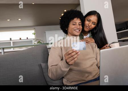 Parcourir le couple diversifié à l'aide d'un ordinateur portable et tenant la carte de crédit dans le salon, avec tasse en céramique Banque D'Images