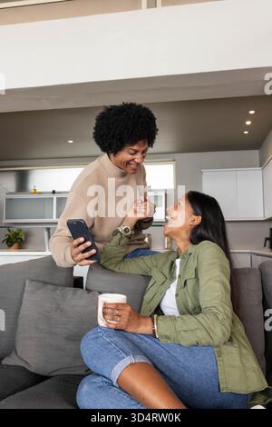 Rire couple diversifié assis sur un canapé gris dans un espace ouvert, avec smartphone et tasse à café Banque D'Images