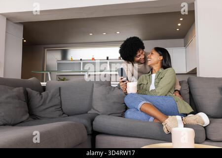 Divers partenaires féminins partageant le café et le smartphone sur le canapé dans le salon moderne Banque D'Images