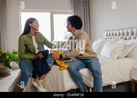 Couple diversifié triant le linge dans une chambre moderne, avec un pull jaune et des vêtements sombres Banque D'Images