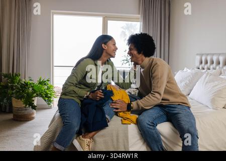 Tri de divers couples manipulant des vêtements pliés sur le lit dans une chambre éclairée, avec des plantes en pot Banque D'Images