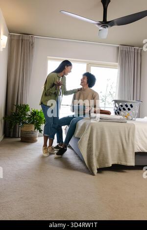 Couple diversifié triant le linge par la fenêtre de la chambre ensoleillée, avec panier à linge et serviettes pliées Banque D'Images