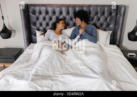 Tenant les mains, couple diversifié couché sous une couette blanche dans une chambre moderne, avec tête de lit touffetée Banque D'Images