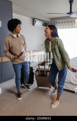 Couple souriant debout dans une chambre moderne, tenant valise et smartphone avec écouteurs Banque D'Images