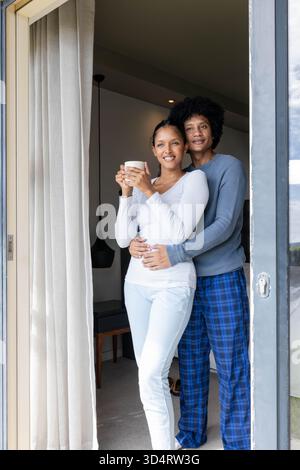 Souriant couple diversifié embrassant à l'intérieur de la pièce doucement éclairée par porte coulissante en verre, tenant tasse en céramique Banque D'Images