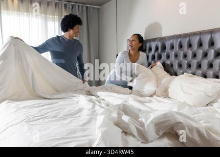 Couple diversifié faisant lit avec draps et oreillers dans une chambre moderne, avec tête de lit touffetée Banque D'Images