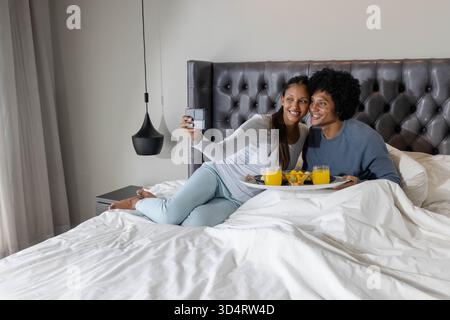 Prise selfie couple diversifié couché sur lit blanc, avec plateau de petit déjeuner et smartphone Banque D'Images
