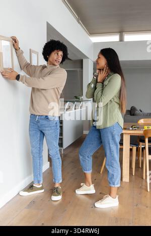 Accrocher et ajuster le cadre photo, couple diversifié décorant le mur du salon, avec ruban à mesurer Banque D'Images