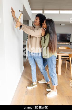 Tenant le marteau couple diversifié fixant le petit clou sur le mur dans le salon décloisonné, avec la lumière du jour Banque D'Images