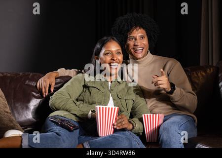 Rire couple diversifié assis sur le canapé dans le salon, tenant des seaux de pop-corn rayés et télécommande Banque D'Images