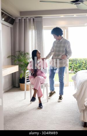 Homme souriant debout à côté d'une femme indienne asiatique assise dans la chambre, avec chaise blanche Banque D'Images