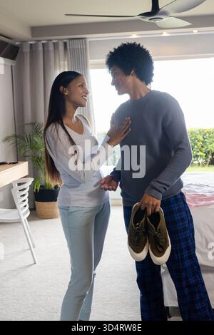 Tenant des baskets, couple diversifié debout dans une chambre ensoleillée, avec lit blanc et plante en pot Banque D'Images