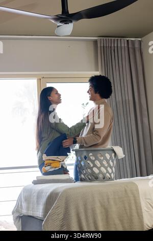 Sourire couple diversifié triant le linge plié sur le lit dans une chambre moderne, avec panier à linge Banque D'Images
