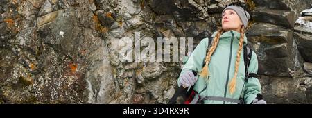 Jeune femme adulte caucasienne avec de longues tresses blondes debout à l'extérieur près d'une falaise rocheuse, tenant des sangles de sac à dos, regardant vers le haut avec une expression réfléchie, portant des équipements de plein air Banque D'Images