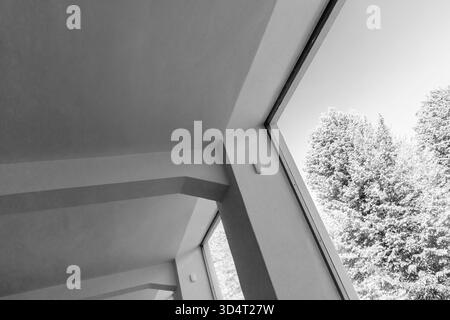 Une scène intérieure monochrome sombre avec des murs inclinés et de grandes fenêtres qui encadrent des arbres feuillus à l'extérieur, créant des lignes épurées, de la lumière naturelle, etc Banque D'Images