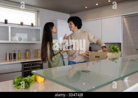 Souriant couple diversifié déballer l'épicerie de la boîte à l'îlot de cuisine, avec smartphone et herbes Banque D'Images