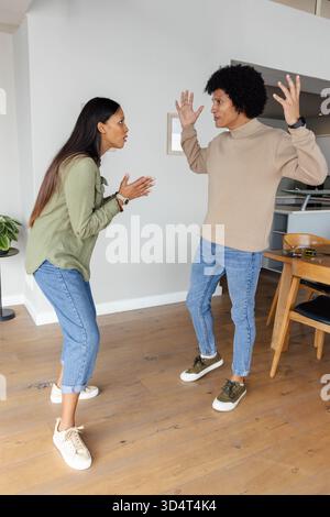 Argumentant couple divers debout face à face dans la maison moderne, avec smartphone sur la table à manger Banque D'Images