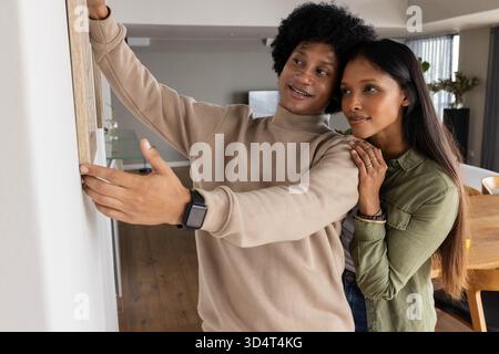 Couple diversifié accrochant l'image encadrée en bois sur le mur dans le salon décloisonné avec des plantes et des stores Banque D'Images