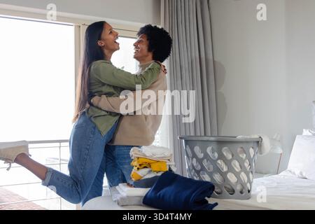Embrasser un couple diversifié souriant dans la chambre près de la porte vitrée coulissante, avec panier à linge, espace copie Banque D'Images