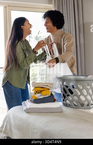 Rire divers couple pliant linge dans une chambre lumineuse, avec panier à linge gris Banque D'Images