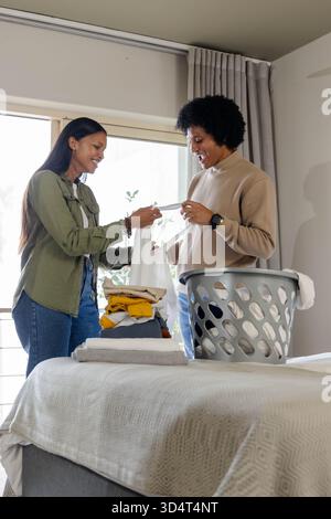 Vêtements pliants divers couple travaillant sur le lit dans une chambre lumineuse, avec panier à linge gris Banque D'Images
