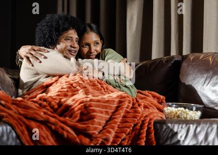 Câlin couple diversifié enveloppé dans une couverture rouge-orange en peluche sur un canapé en cuir à la maison, avec pop-corn Banque D'Images