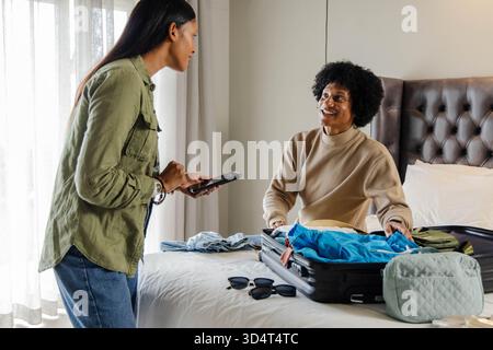 Emballage de couple diversifié remplissant la valise ouverte avec des vêtements sur le lit dans la chambre, tenant smartphone Banque D'Images