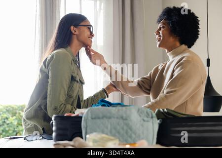 Homme souriant et femme indienne échangeant contact tout en emballant des articles à la maison, avec valise noire ouverte Banque D'Images