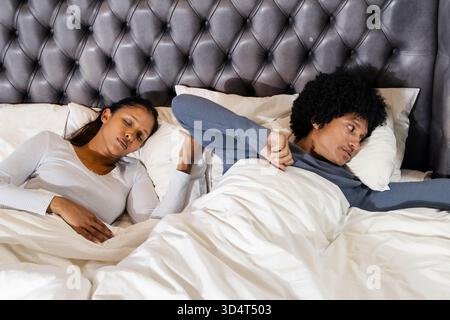 Couple de repos diversifié sur le lit sous couette dans la chambre, montrant des oreillers blancs et la tête de lit Banque D'Images