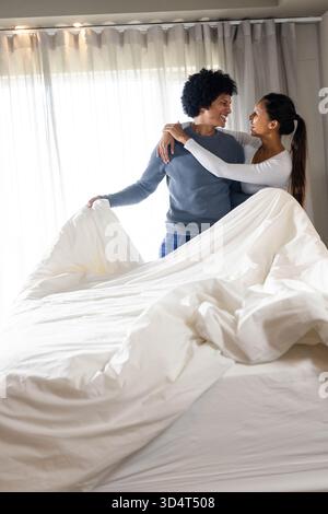 Couple diversifié faisant lit dans une chambre aérée, avec housse de couette blanche et rideaux transparents par fenêtre Banque D'Images