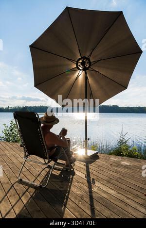 Le dos d'un homme âgé assis dans une chaise longue sur une terrasse en bois au bord d'un lac Banque D'Images