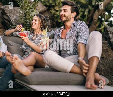 Deux amis s'assoient confortablement sur un élégant canapé extérieur, sirotant des cocktails vibrants. Le rire remplit l'air alors qu'ils se détendent parmi la verdure luxuriante sous un cle Banque D'Images