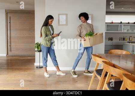 Couple diversifié debout dans l'entrée ouverte de la maison, tenant la boîte de fruits et de légumes et vérifiant le smartphone Banque D'Images