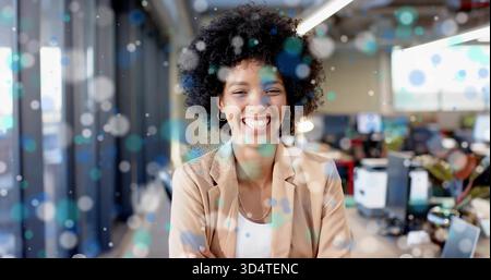 Femme souriante portant blazer beige et collier debout dans un bureau moderne, avec des moniteurs et des plantes Banque D'Images