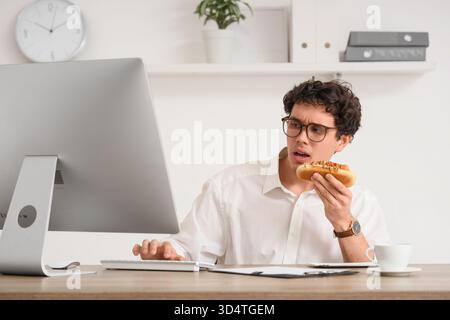 Jeune homme d'affaires avec hot dog savoureux travaillant dans le bureau Banque D'Images
